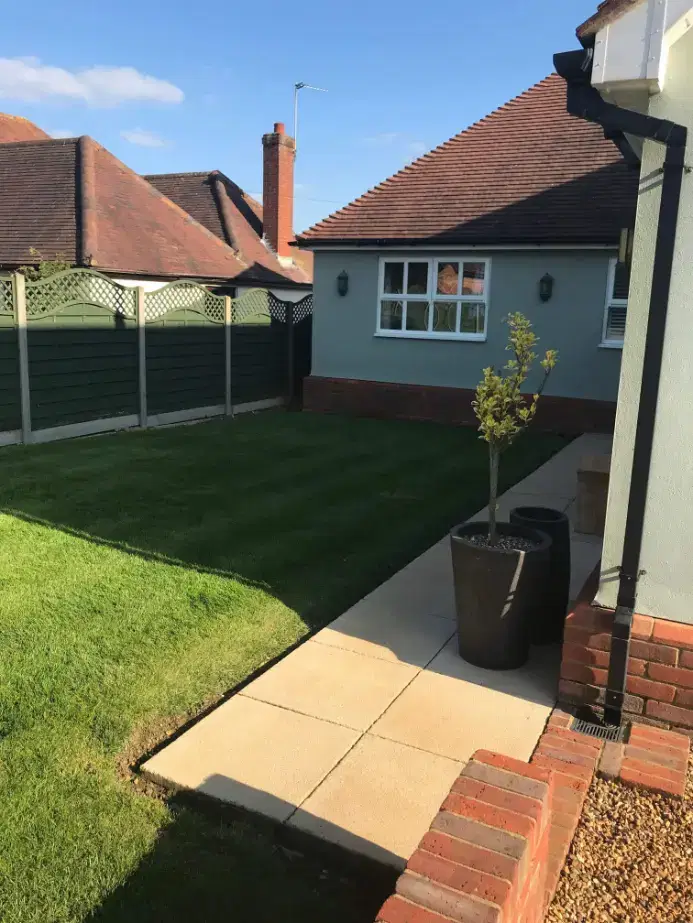 Backyard lawn with a paved walkway, two tall black plant pots, a small tree, and houses with red roofs and fences.